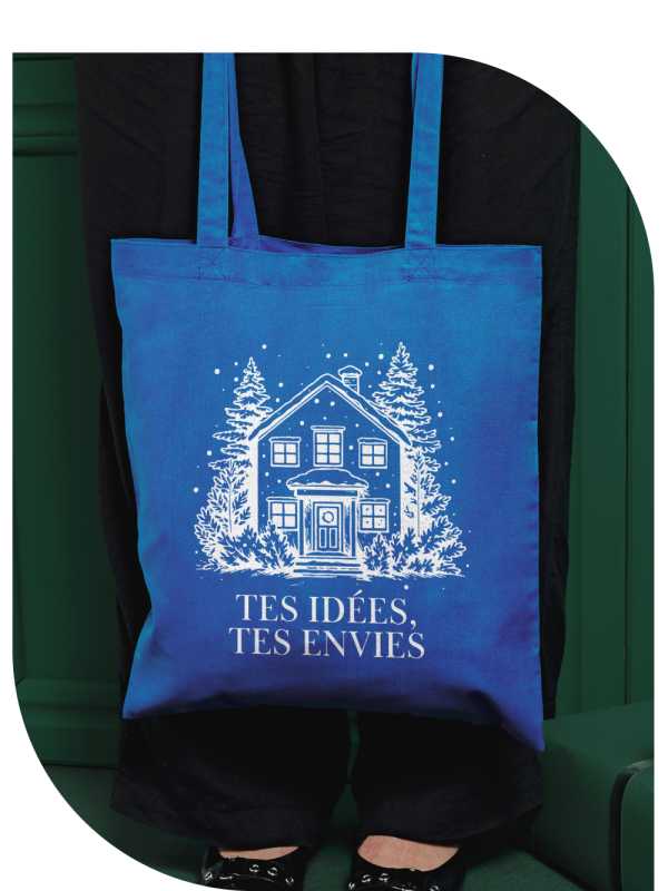 Sac bleu avec un design de maison blanche et le texte « Ton idée, ton envie », tenu devant un pantalon foncé et un fond vert.
