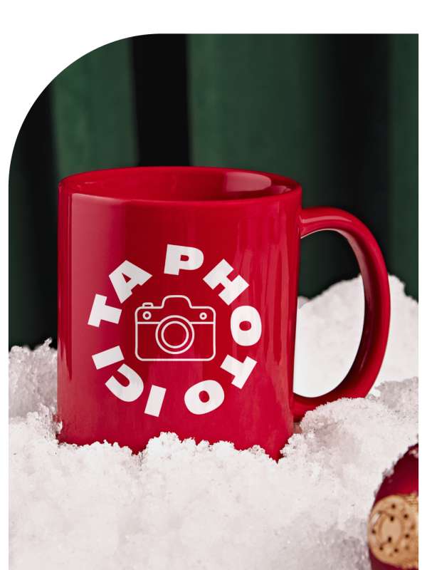 Tasse rouge avec une icône de caméra blanche et le texte « Ta photo ici », posée dans la neige devant un fond vert.