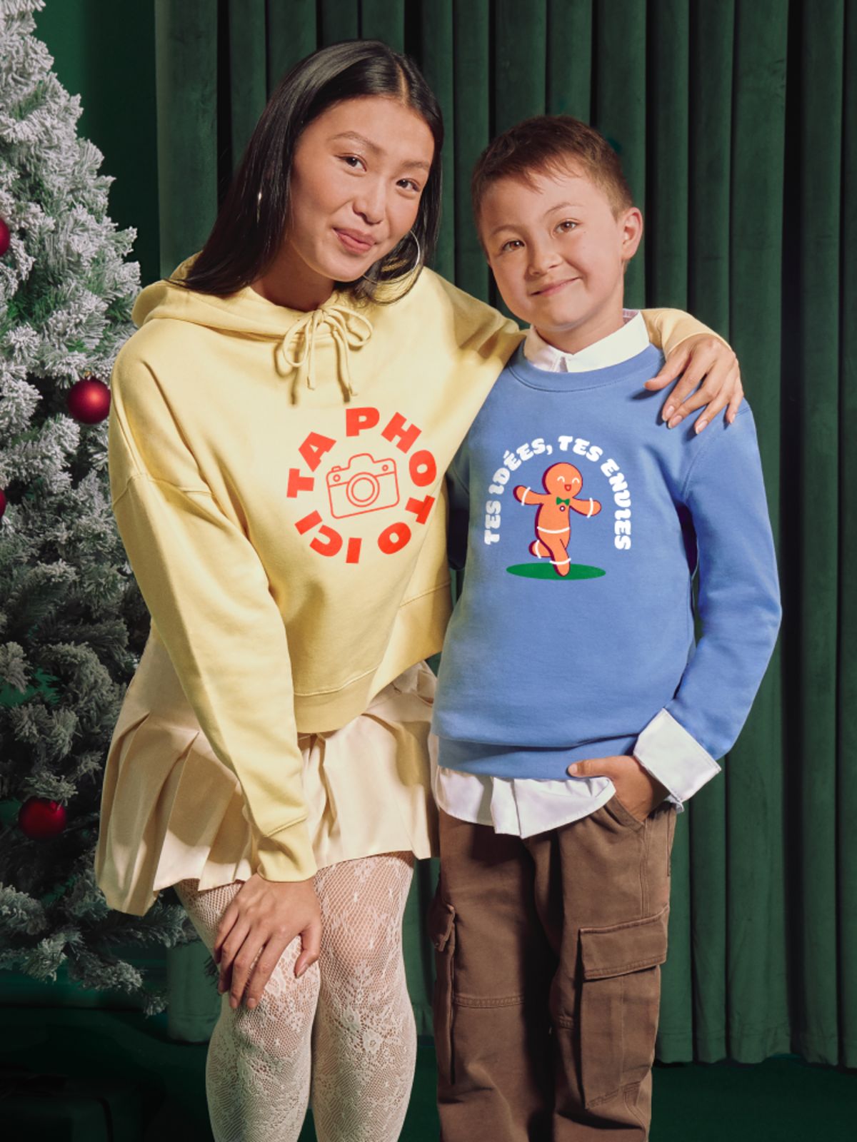 Three people wearing personalized christmas hoodies in front of a green curtain and decorated tree.