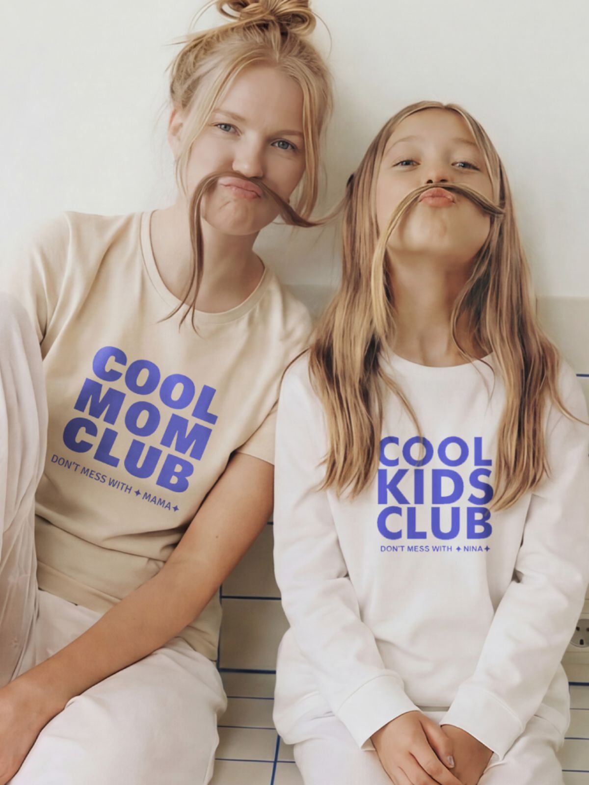 Mother and daughter pose against a blue-and-white tiled wall, wearing tops with “Cool Mom Club” and “Cool Kids Club”.