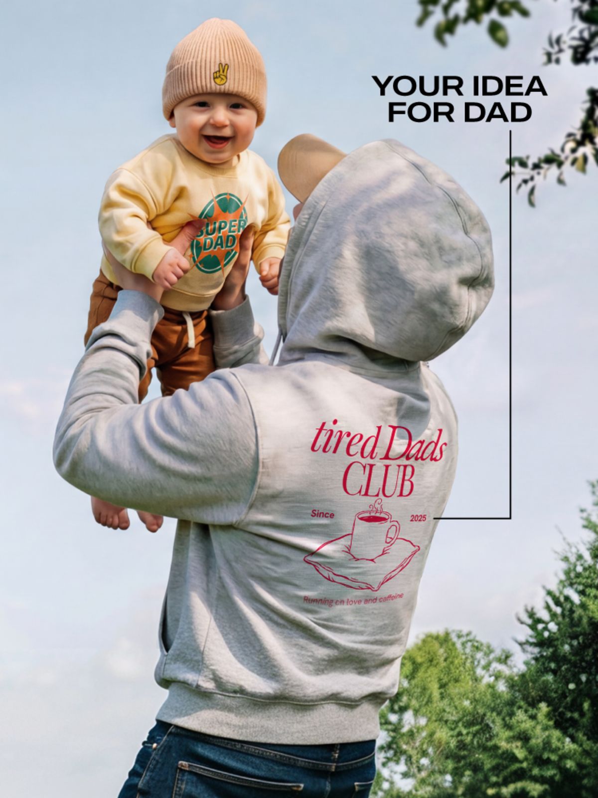 Father lifts smiling baby outdoors wearing hoodie with 'tired Dads Club' print and text 'Your idea for dad'.