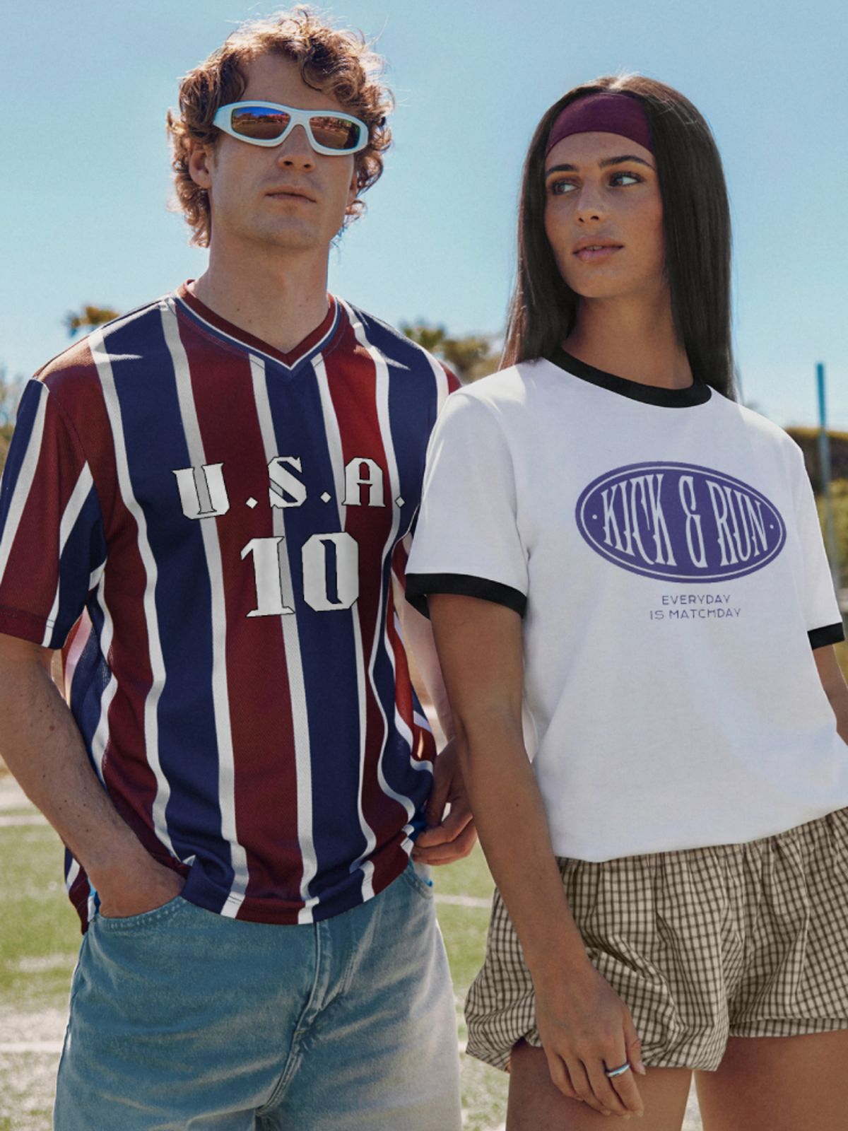 Two people stand on a football field wearing printed retro shirts while a football match is played in the background.
